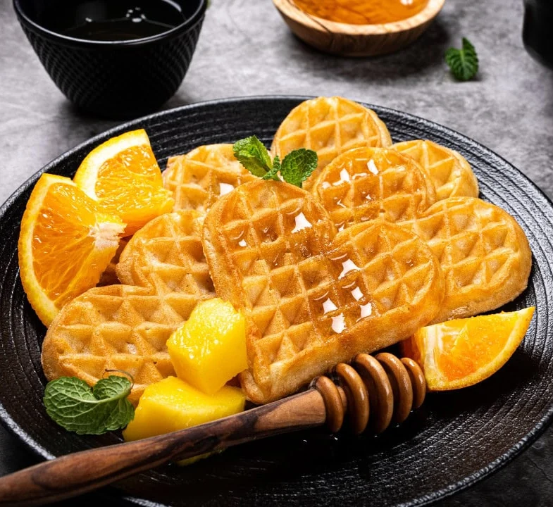 Herzwaffeln mit Honig, Orangenspalten und Mango auf einem schwarzen Teller
